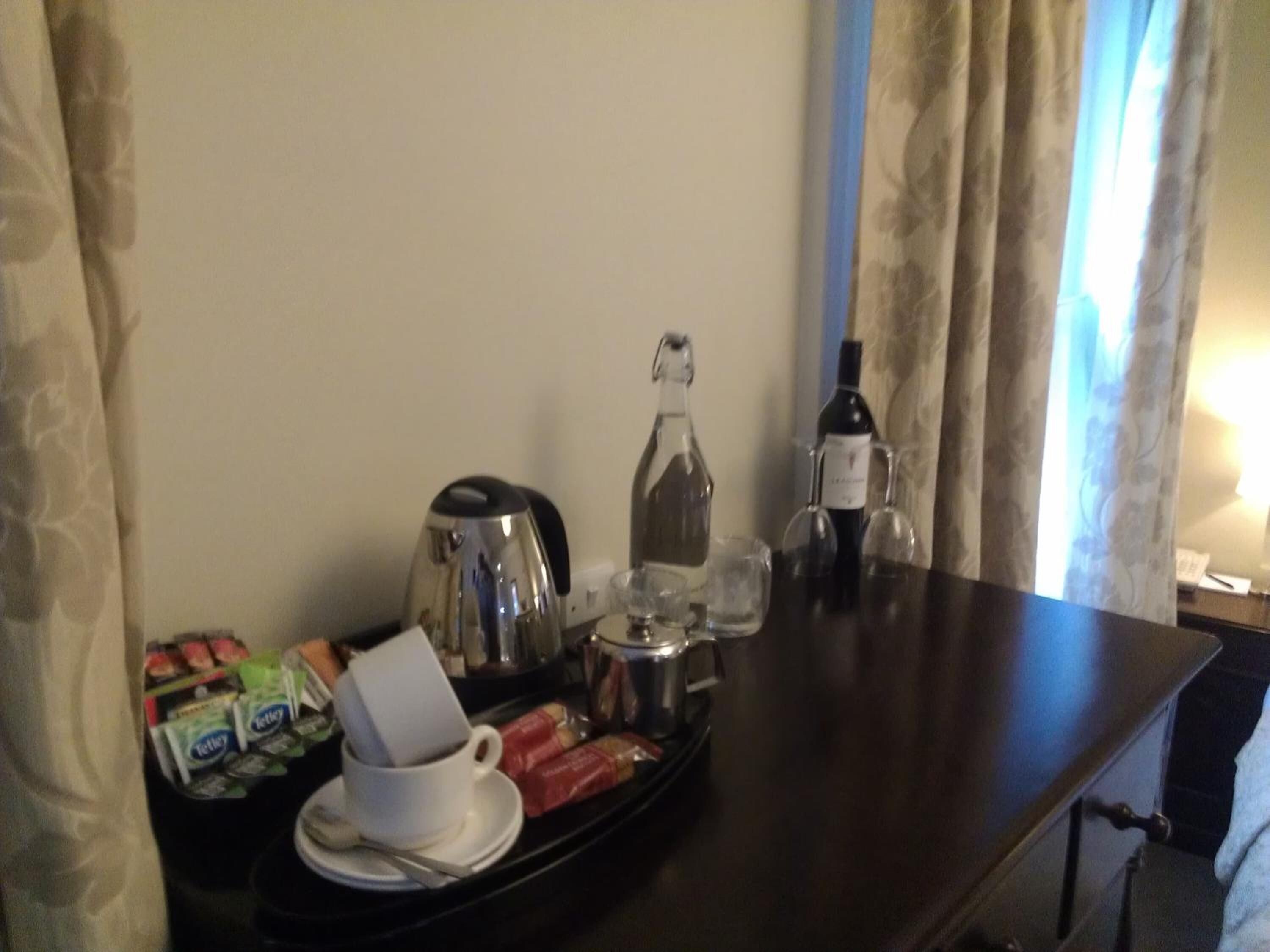 A hotel room with a dining table set with a coffee pot, a bottle of wine, a glass, a cup, and some packaged food.