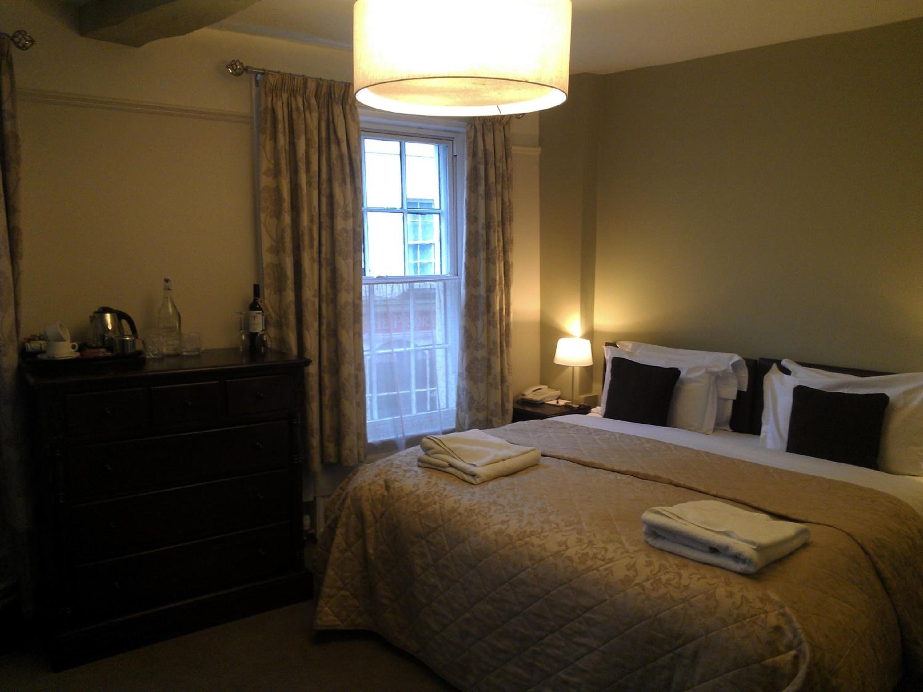 Una habitación de hotel con cama, cómoda, mesita de noche, lámpara y ventana con cortinas.