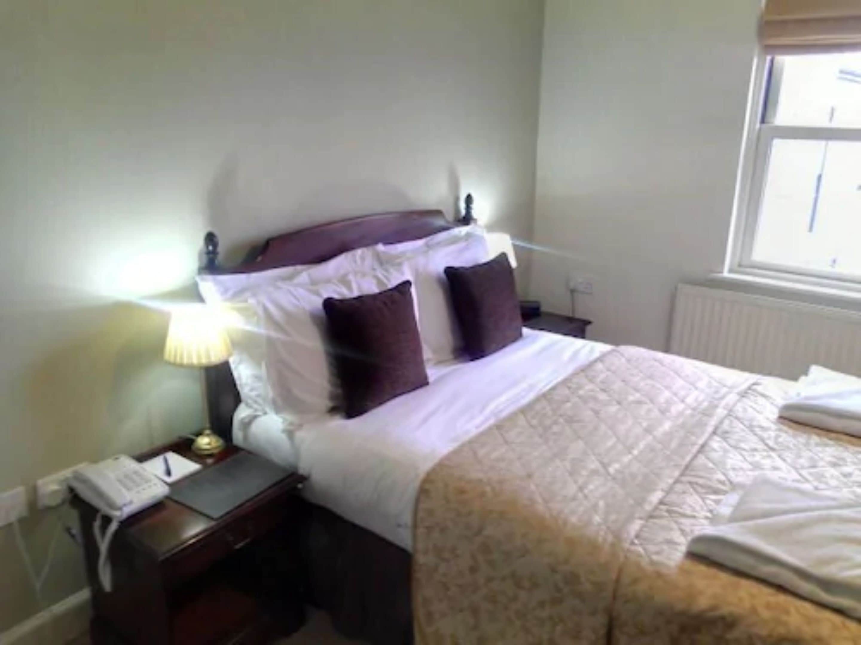A neatly made bed with a white comforter and two dark pillows, a bedside lamp, and a small wooden nightstand with a telephone and a book.