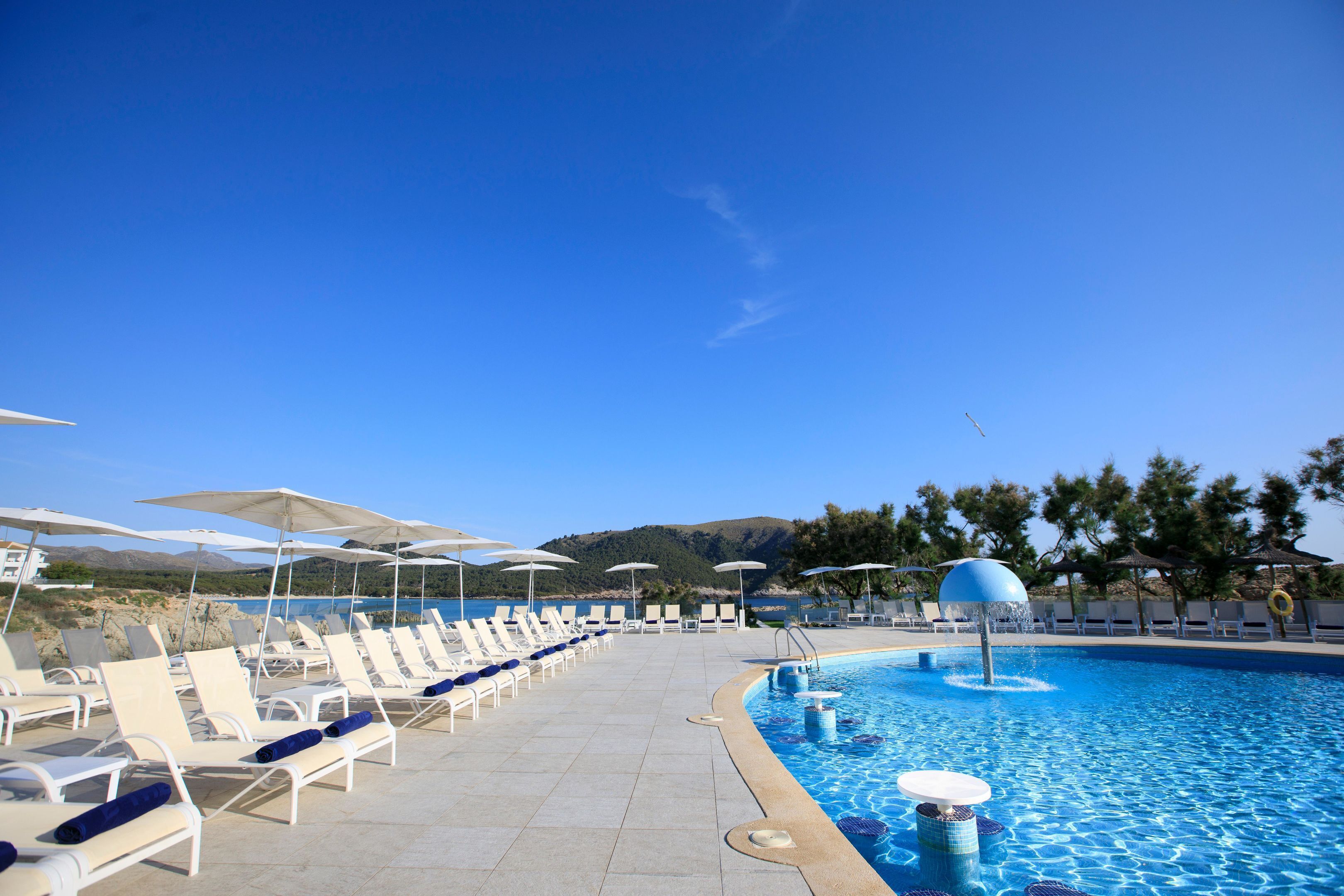 Un área junto a la piscina con sillones de descanso y sombrillas, un cielo azul despejado y montañas al fondo.