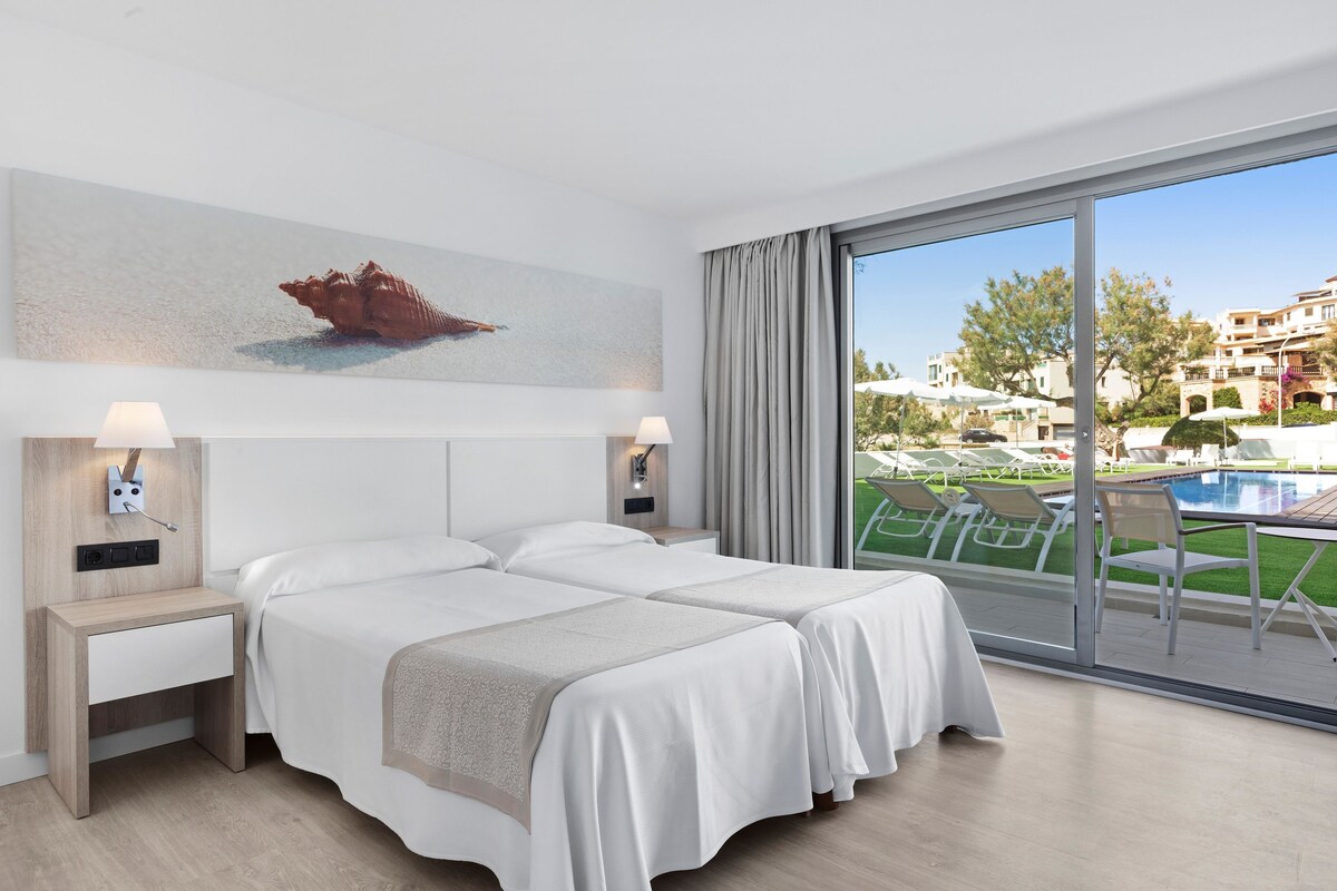 A hotel room with two beds, a large window with a view of a pool and buildings, and a painting on the wall.