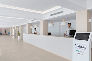 Zona de recepción de hotel moderna con un mostrador blanco, un quiosco digital y una recepcionista.
