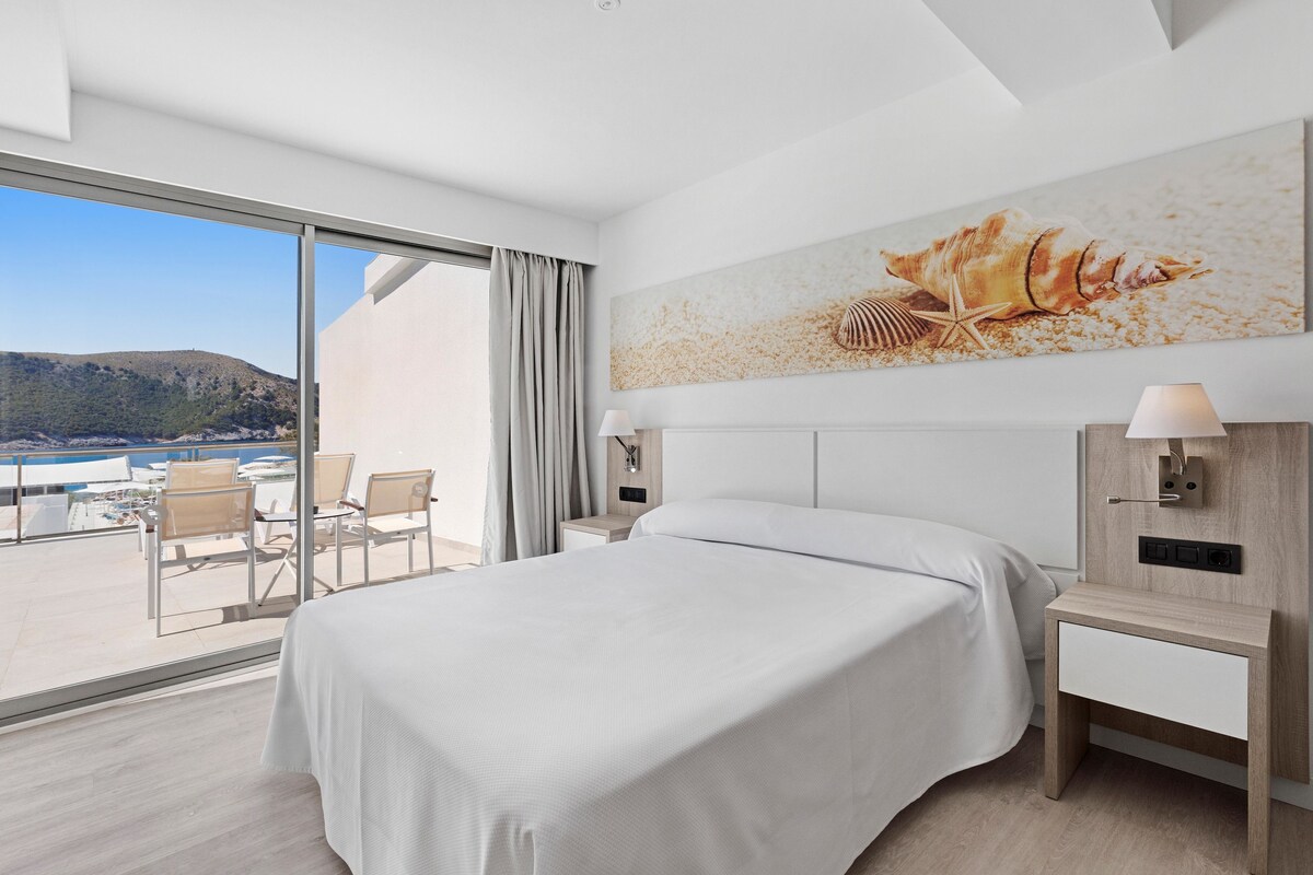 A hotel room with a large bed, a desk, a chair, a view of the sea, and a painting of seashells.