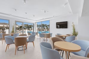 A modern indoor seating area with tables and chairs, large windows, and a pool view. - THB Cala Lliteras Hotel - Adults Only (Capdepera)