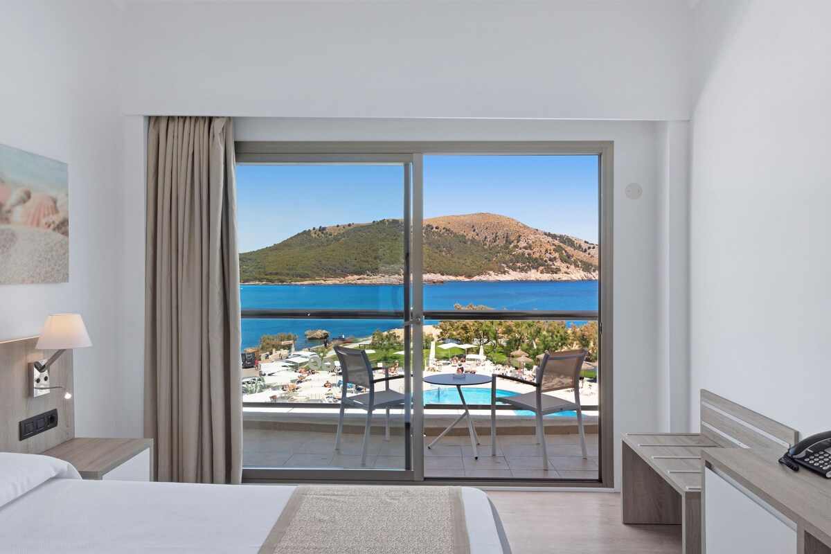 A hotel room with a bed, a desk, a chair, and a view of a pool and beach.