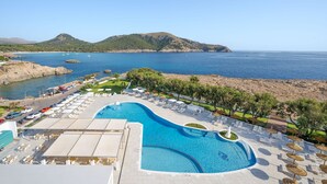Ein Resort mit einem großen Schwimmbad, Liegestühlen und Blick auf das Meer und die Hügel.