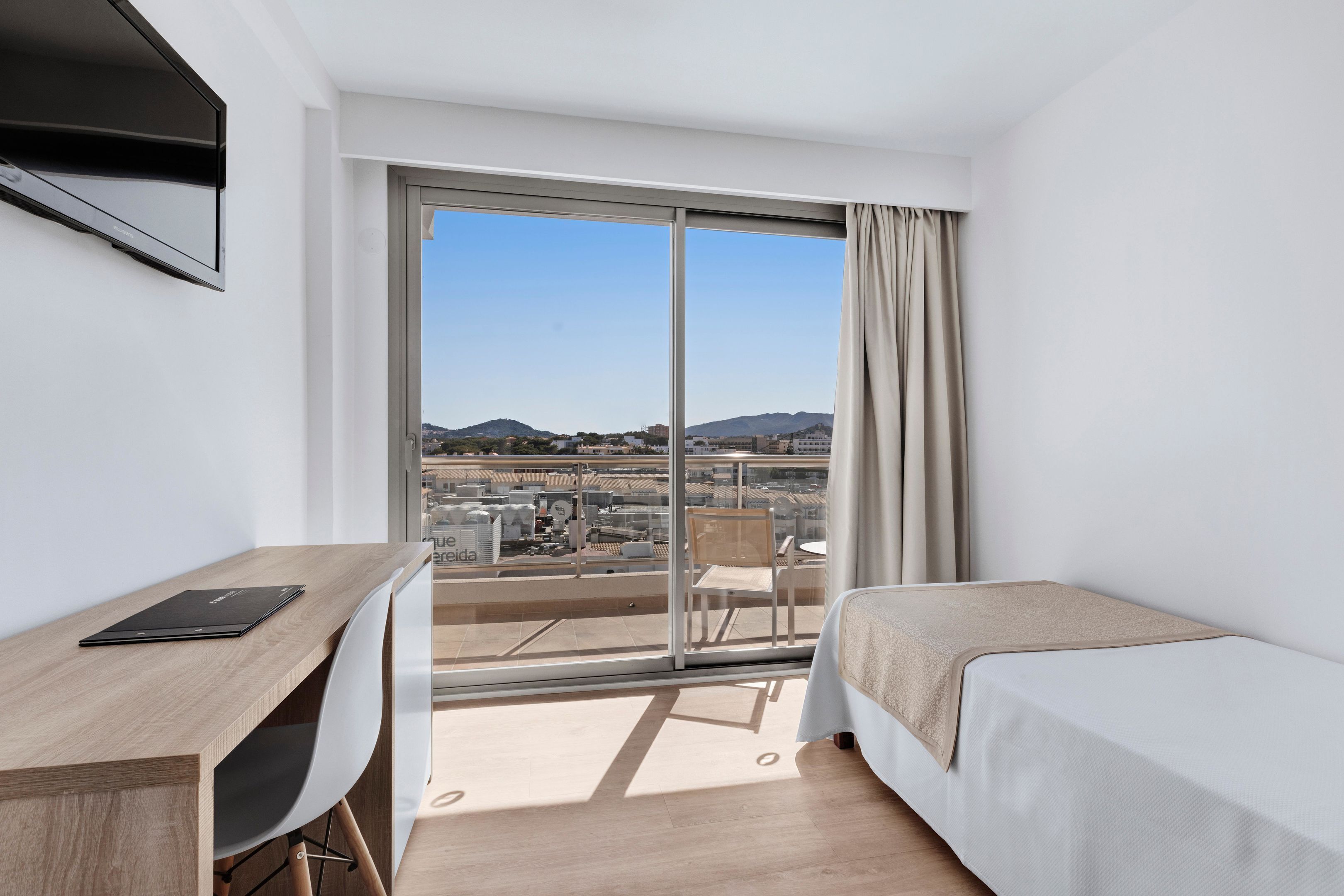 Habitación de hotel con cama, escritorio con una computadora portátil, una silla y un amplio ventanal con vistas a la ciudad.