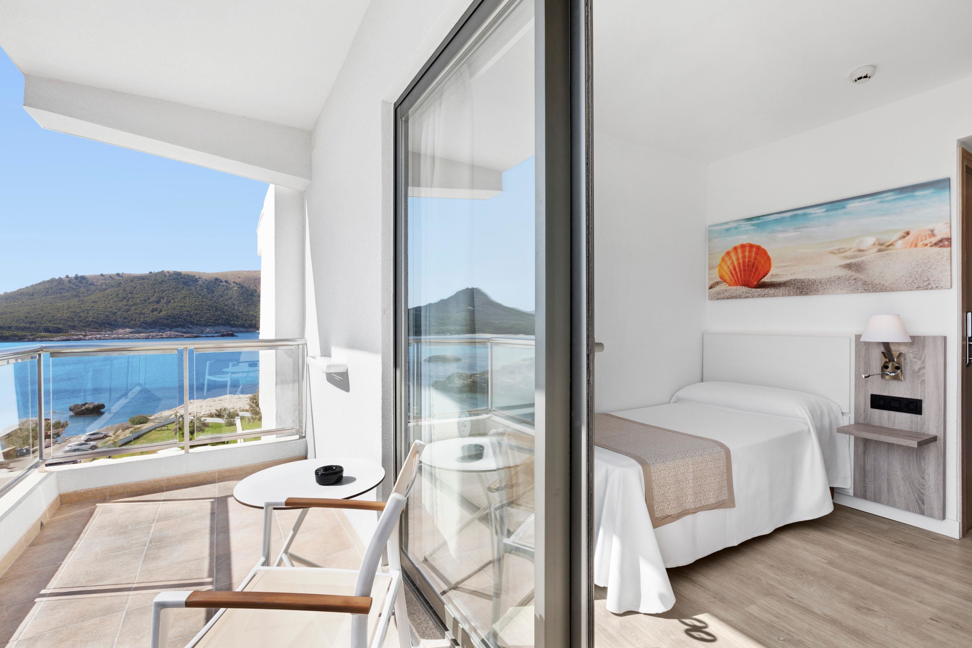 Habitación de hotel moderna con balcón y vista panorámica de la costa, una cama, una mesita y un cuadro de una playa.