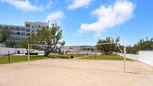 Una cancha de voleibol de arena con una red, rodeada de árboles y un edificio al fondo.