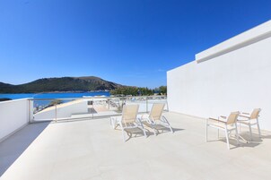 A rooftop terrace with white chairs and a view of the sea and hills.