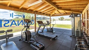 Un gimnasio cubierto con equipamiento de ejercicio, vista al exterior y un cartel con texto parcialmente visible.