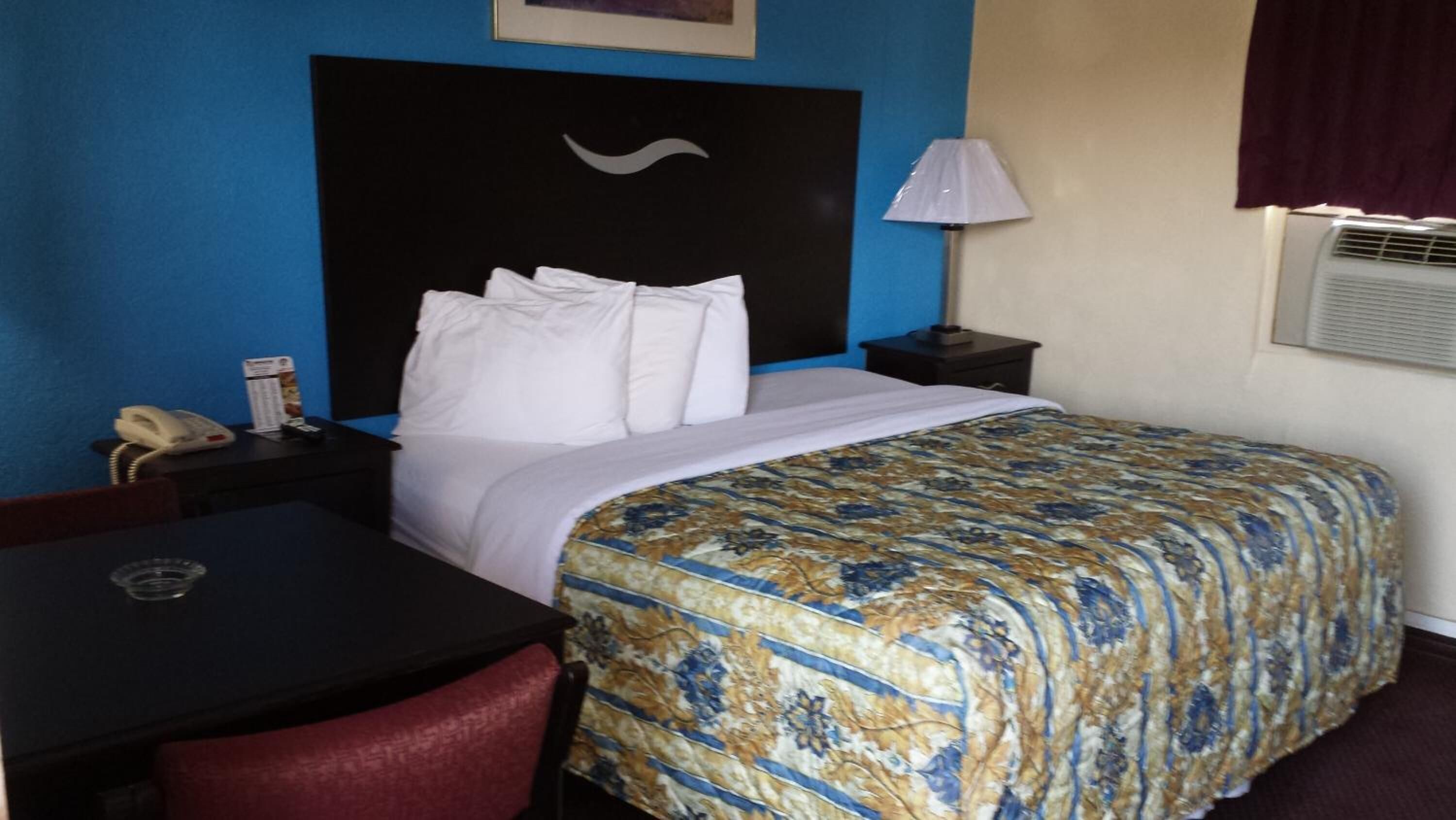 A hotel room with a bed, a desk, a chair, a phone, a lamp, and an air conditioning unit.