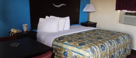 A hotel room with a bed, a desk, a chair, a phone, a lamp, and an air conditioning unit.