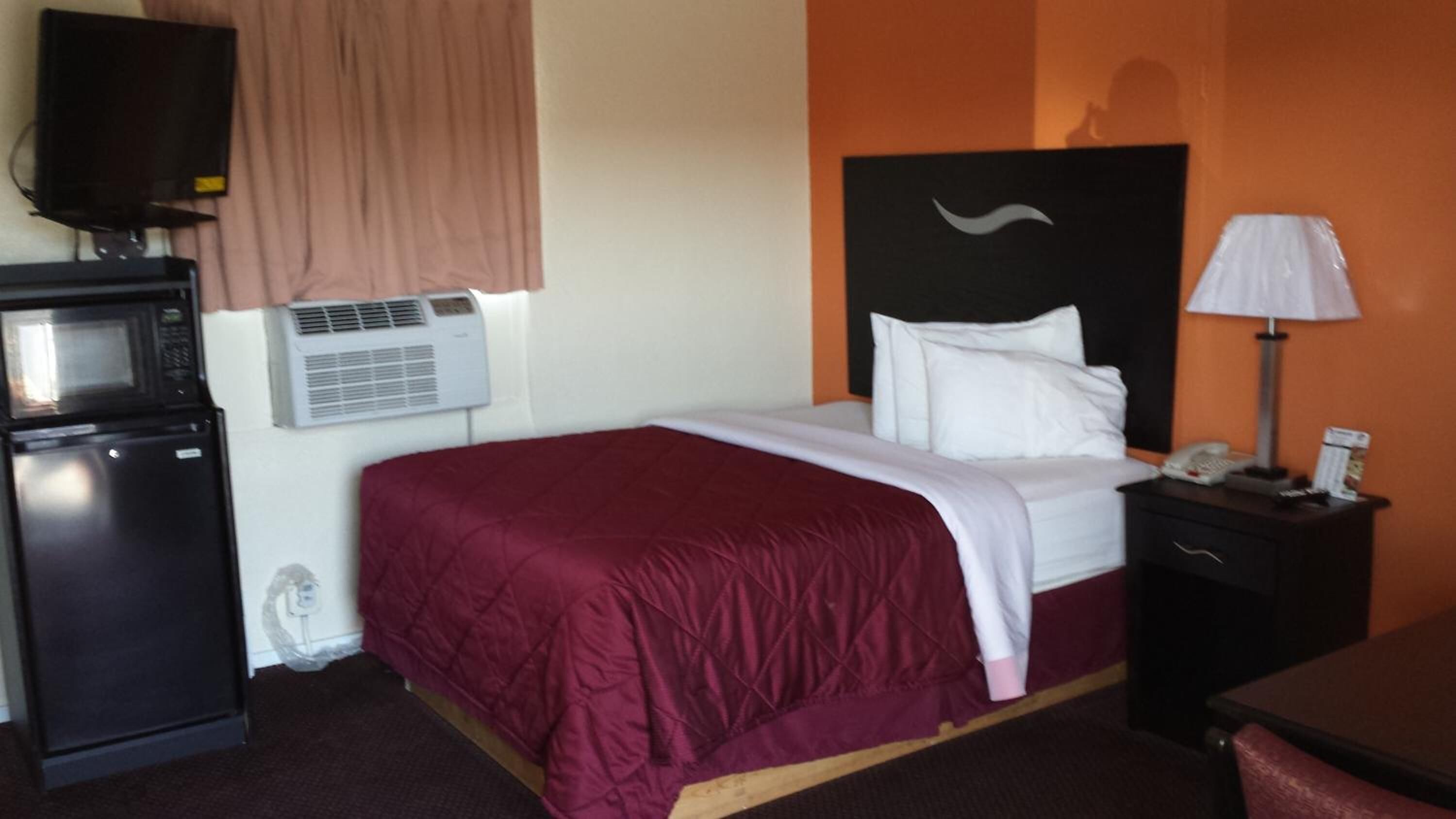 A hotel room with a bed, a desk, a lamp, a telephone, a microwave, and an air conditioner.