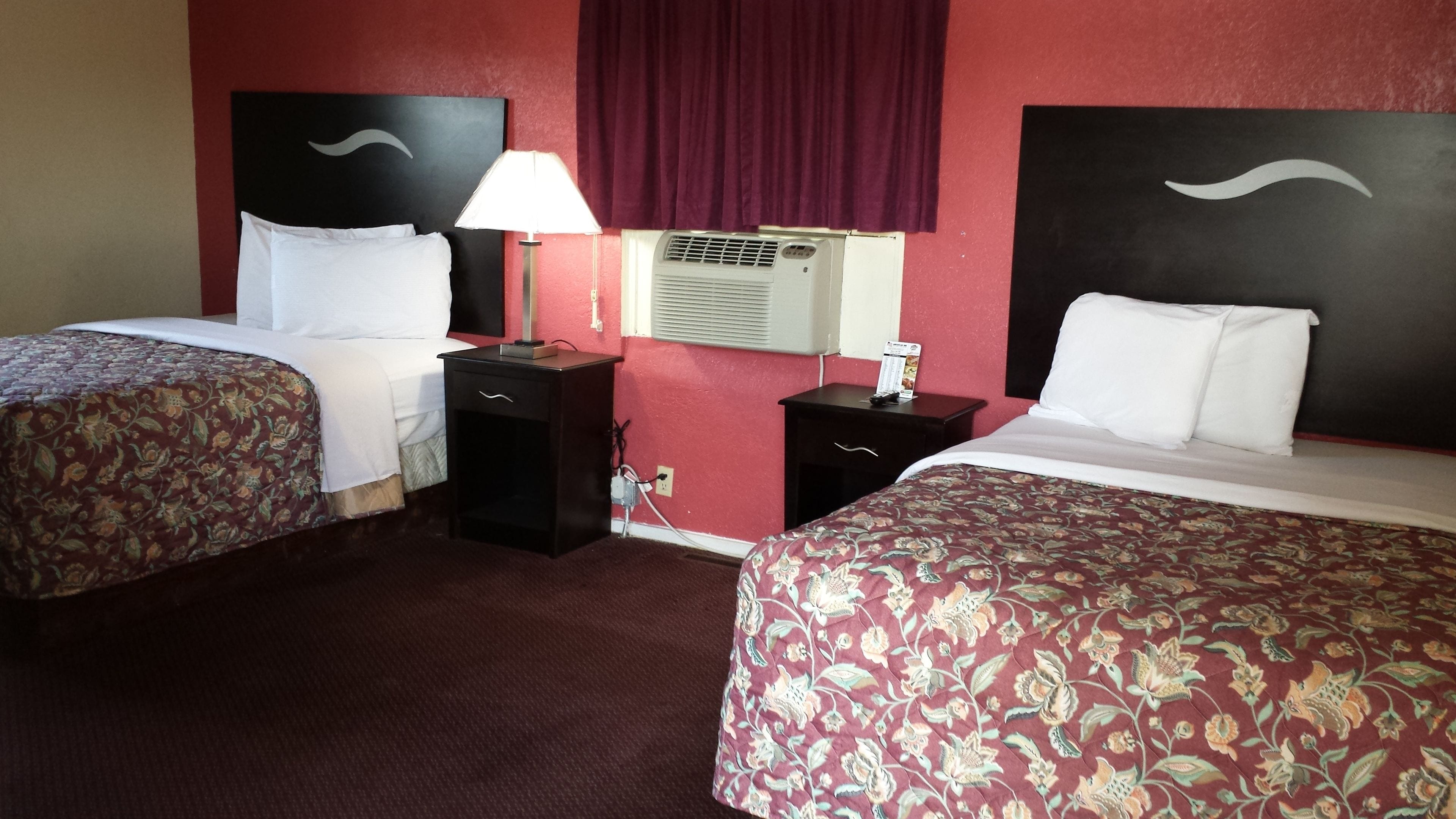 Habitación de hotel con dos camas, una mesita de noche con lámpara, un aire acondicionado y una cortina roja.