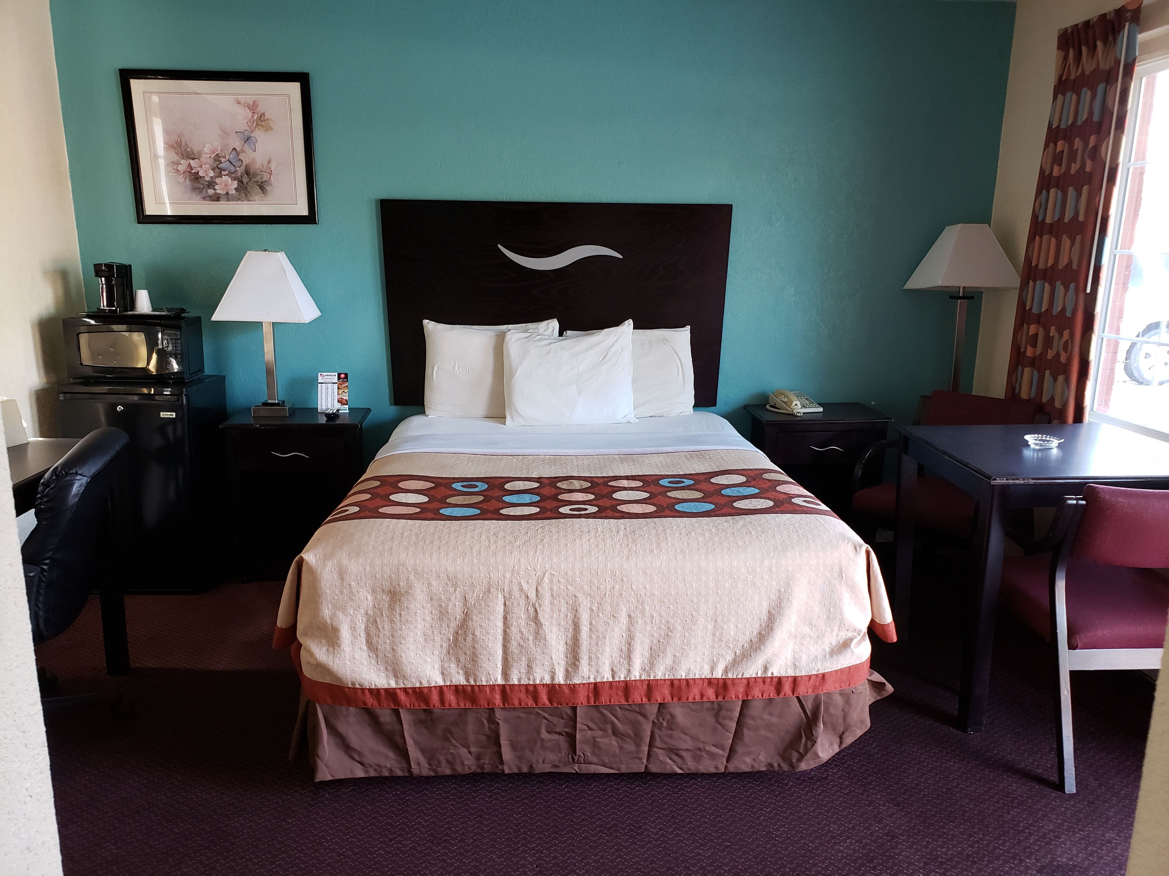 A hotel room with a bed, two bedside tables, a desk, a chair, a microwave, a telephone, and a picture on the wall.