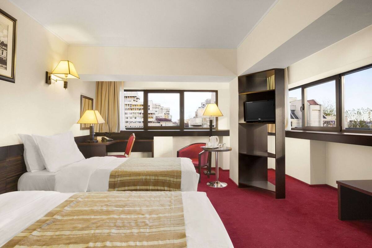 A hotel room with two beds, a desk, a chair, a TV, and a window with a city view.