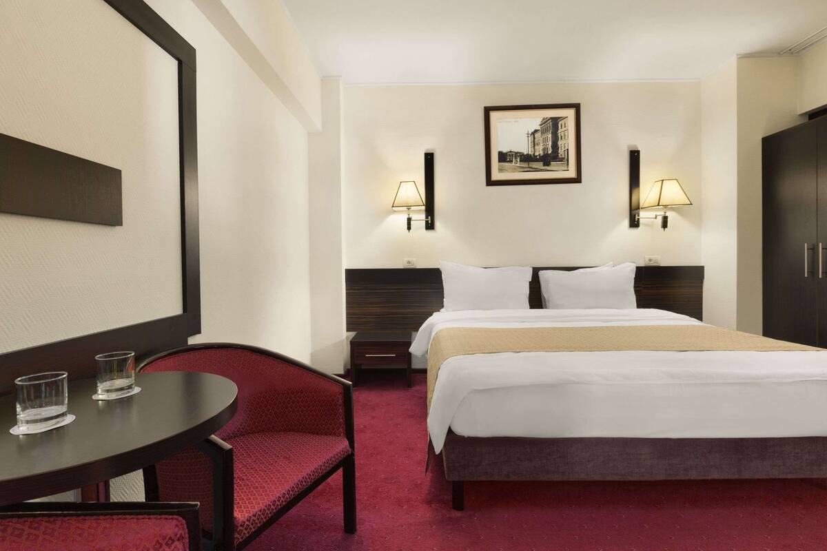 A hotel room with a bed, two bedside tables, a chair, a round table, and a framed picture on the wall.
