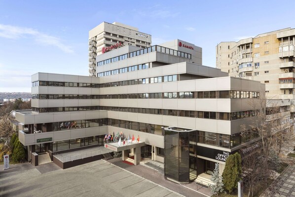 Un edificio moderno de varios pisos con un llamativo letrero de 'RAMADA', que cuenta con una entrada de vidrio y una sección identificada como 'THE OFFICE LOUNGE'.