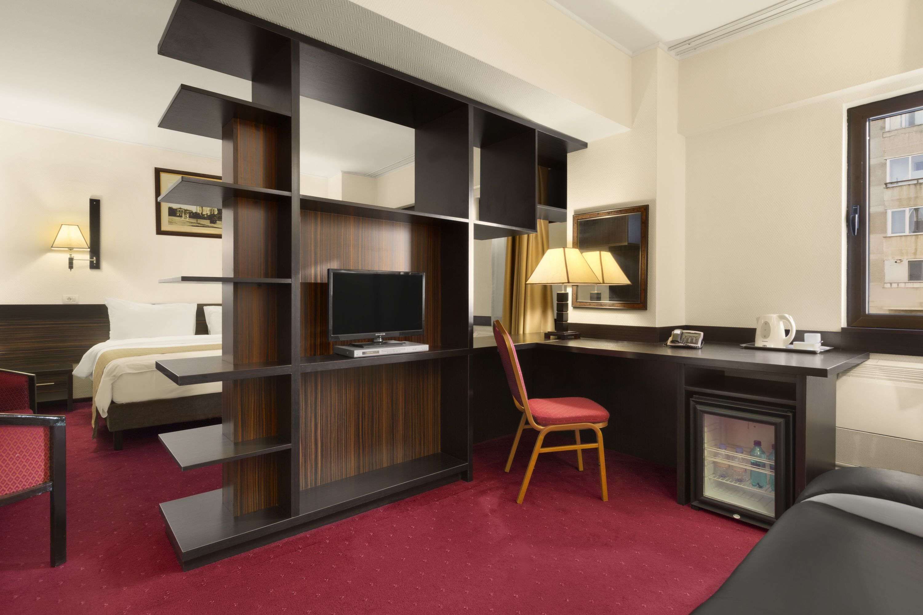 A hotel room with a bed, a desk with a computer, a chair, a television, and a minibar.