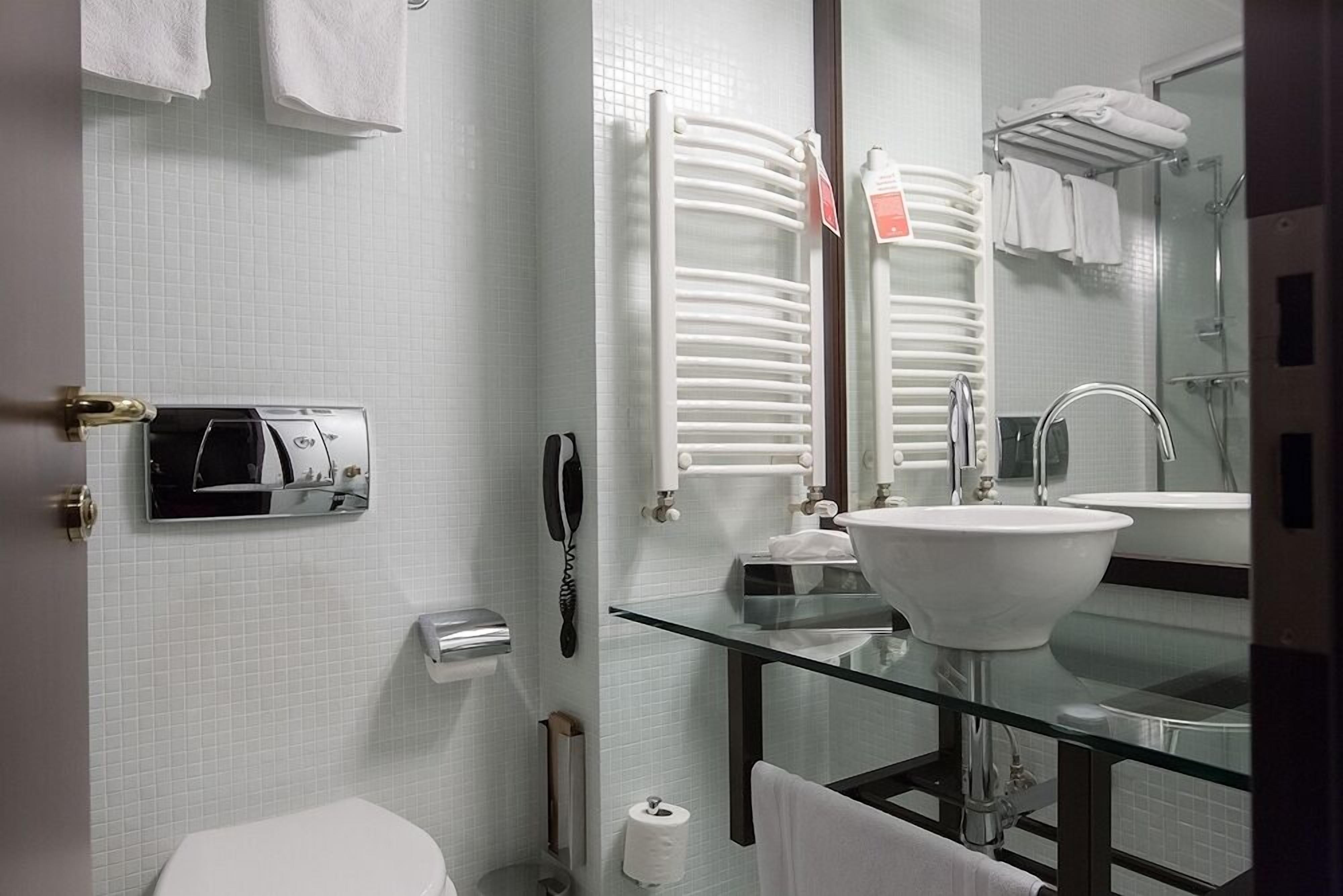Un baño moderno con un lavamanos de vidrio, azulejos blancos, un inodoro, una cabina de ducha con mampara de vidrio y un perchero para toallas.
