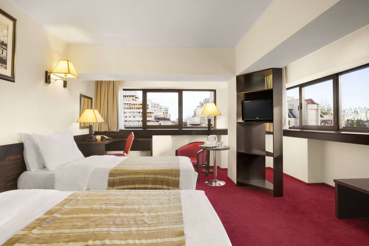 A hotel room with two beds, a TV, a small table, and a window with a city view.