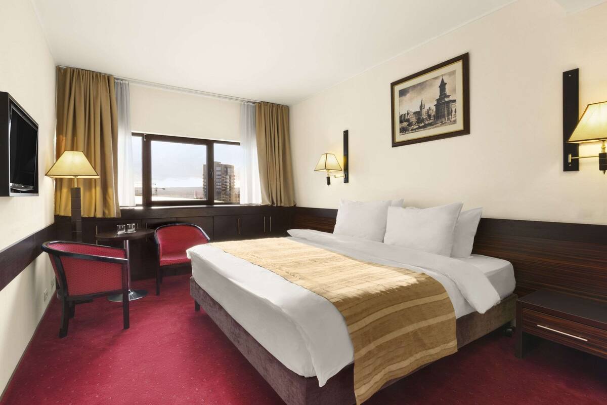 A hotel room with a large bed, a desk, a chair, and a window with a city view.