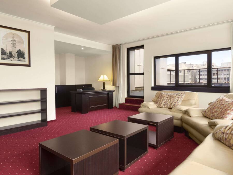 A hotel room with a red carpet, a sofa set, a desk, a lamp, and a large window with a city view.