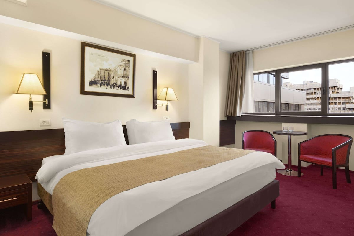 A hotel room with a large bed, two bedside lamps, a framed picture on the wall, a small table with a chair, and a window with a view of buildings.