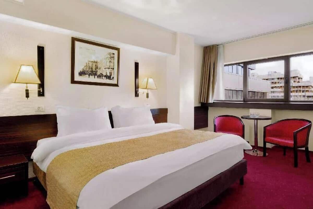A hotel room with a bed, two bedside lamps, a window with a view, and a small table with a chair.