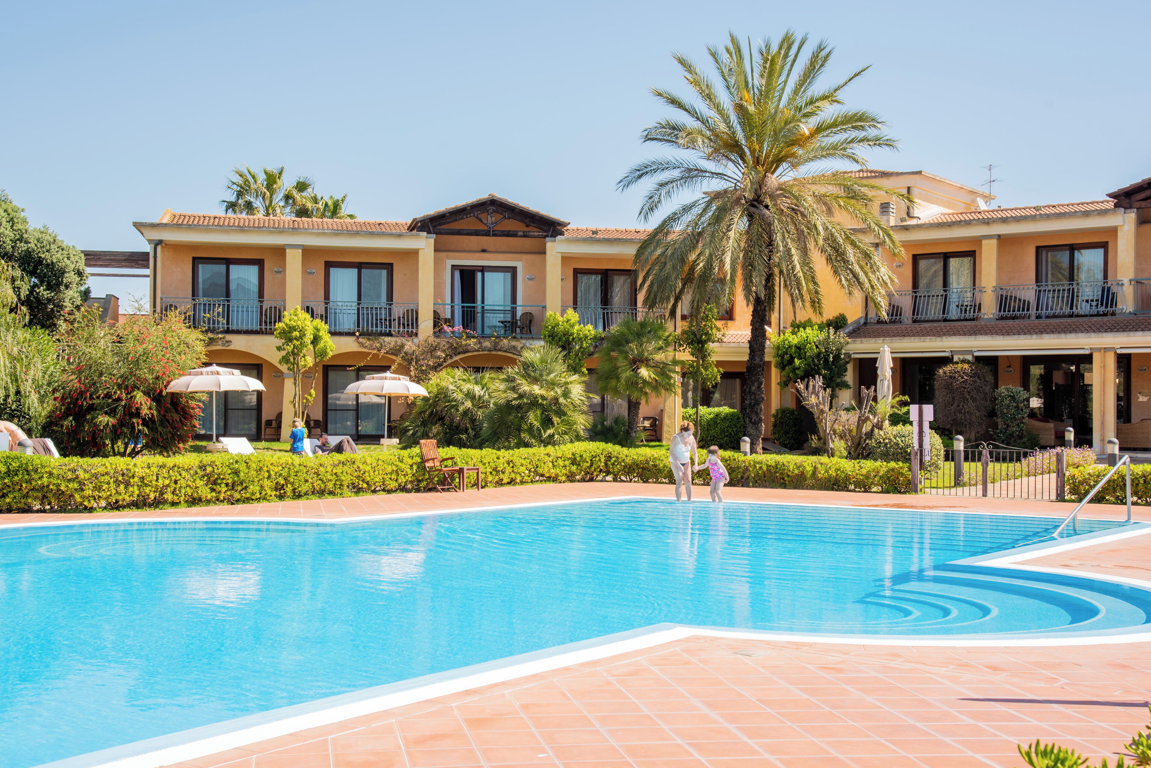 A swimming pool with a tiled deck, a palm tree, and a building with balconies and windows.