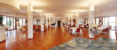 Un amplio vestíbulo de hotel con suelo de baldosas, varias zonas de comedor y grandes ventanales.