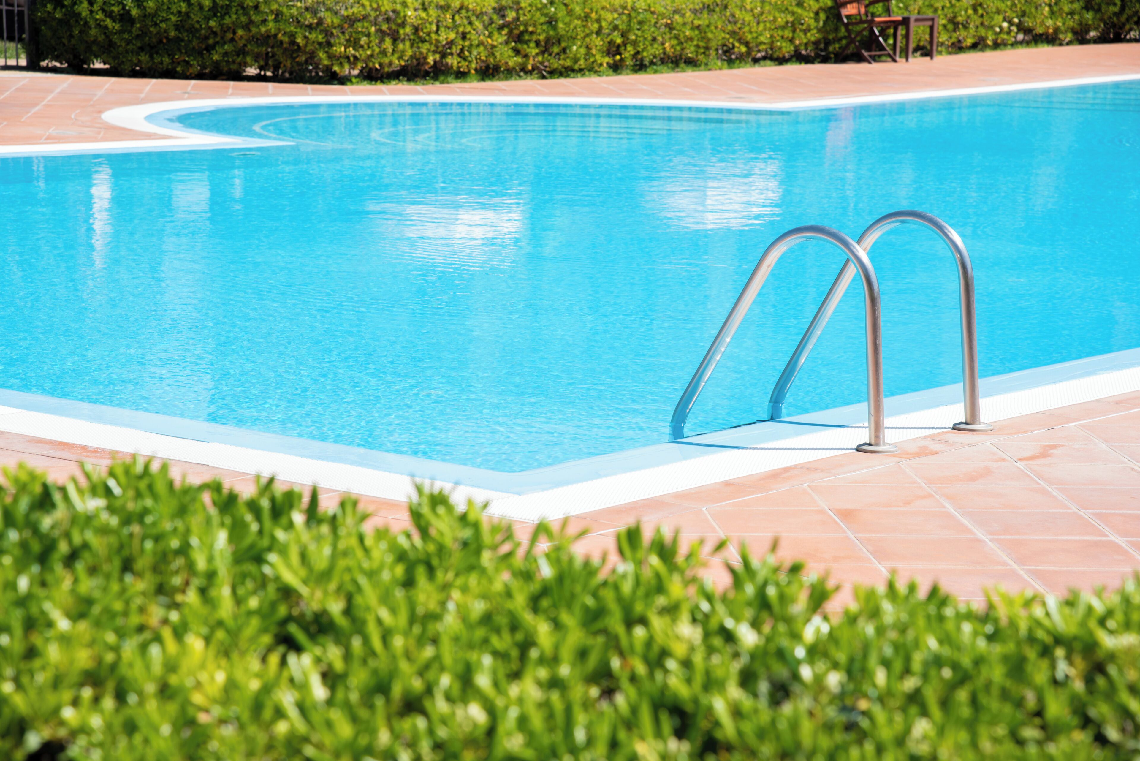 A swimming pool with metal ladders, surrounded by a tiled deck and green bushes.