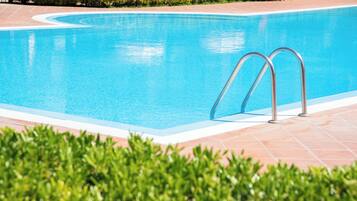 A swimming pool with metal ladders, surrounded by a tiled deck and green bushes.