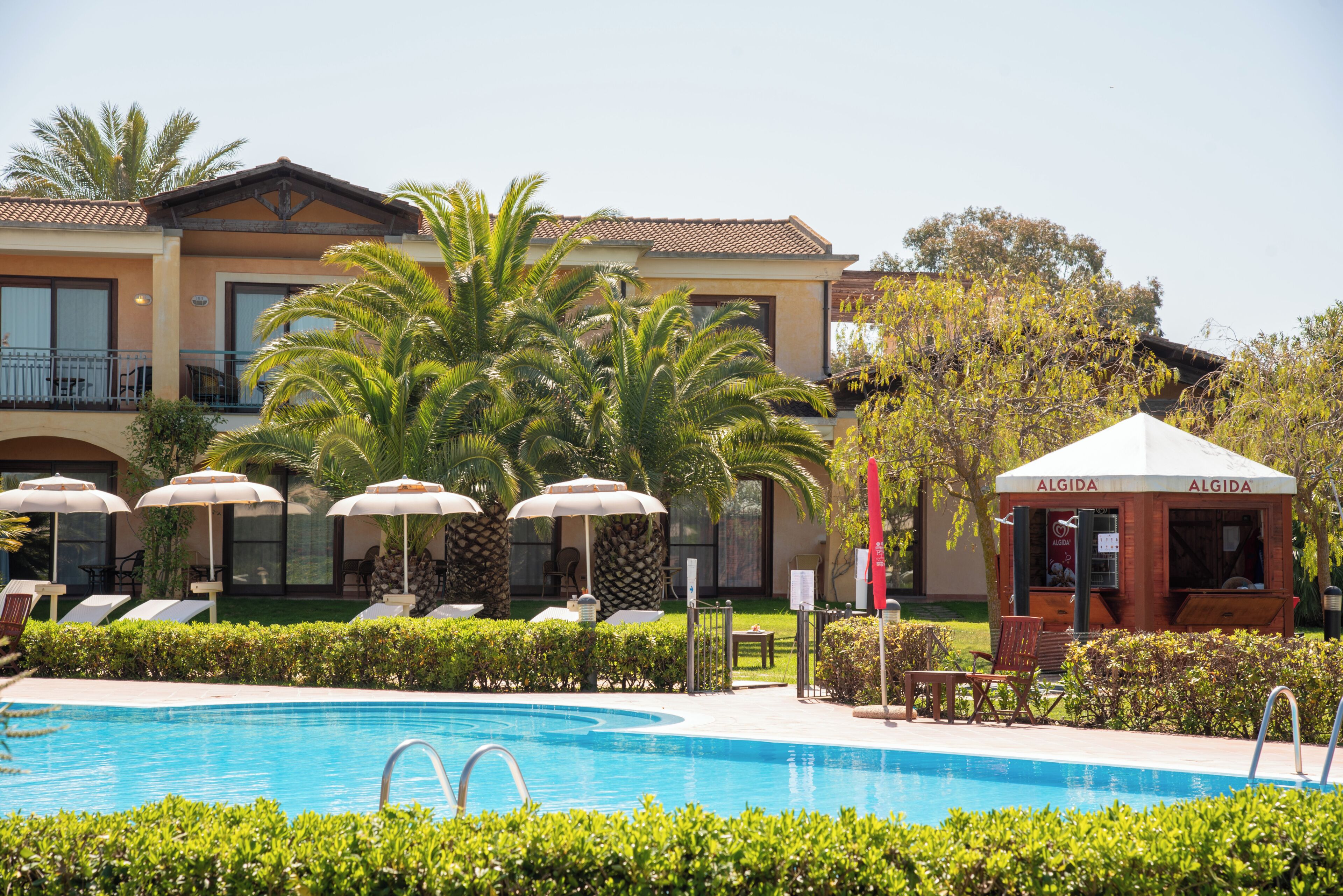 En swimmingpool med liggestole og parasoller, en pergola og en bygning med tagsten.