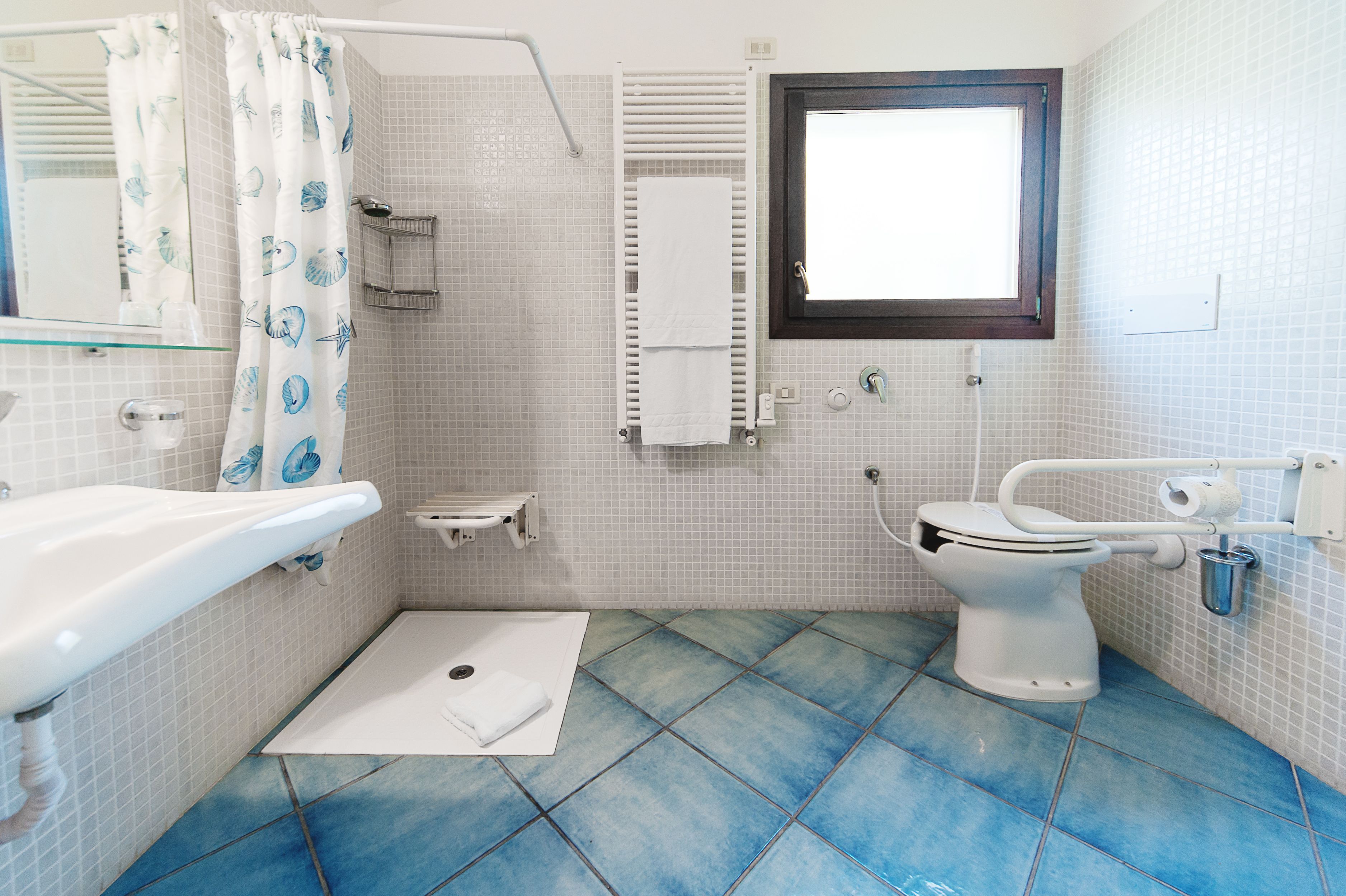 Ein Badezimmer mit weißem Waschbecken, einer Toilette mit Haltegriff, einer Duschkabine mit Vorhang und einem Fenster.