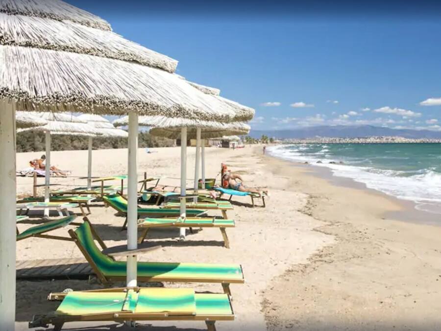 Uma praia com espreguiçadeiras e toldos de palha, areal extenso e vista clara do oceano.