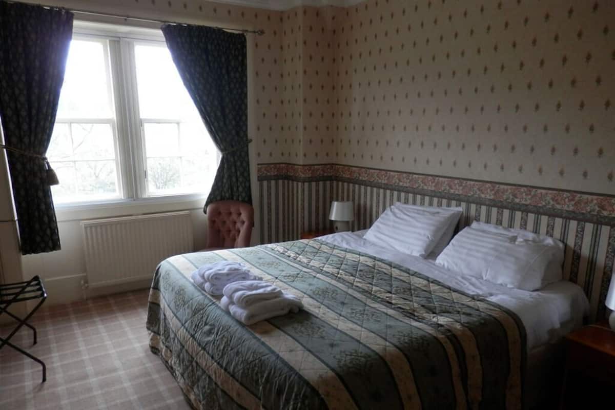 Quarto de hotel com uma cama, um radiador, uma janela com cortinas e uma luminária.