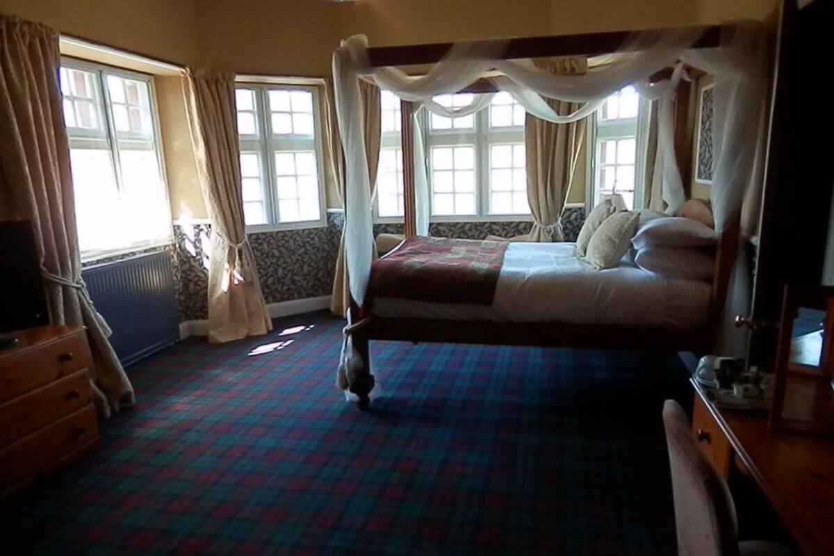 Uma cama com dossel e cortina, um aparador de madeira e um tapete estampado em um quarto com janelas amplas e cortinas.