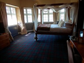 A four-poster bed with a canopy, a wooden dresser, and a patterned carpet in a room with large windows and curtains.