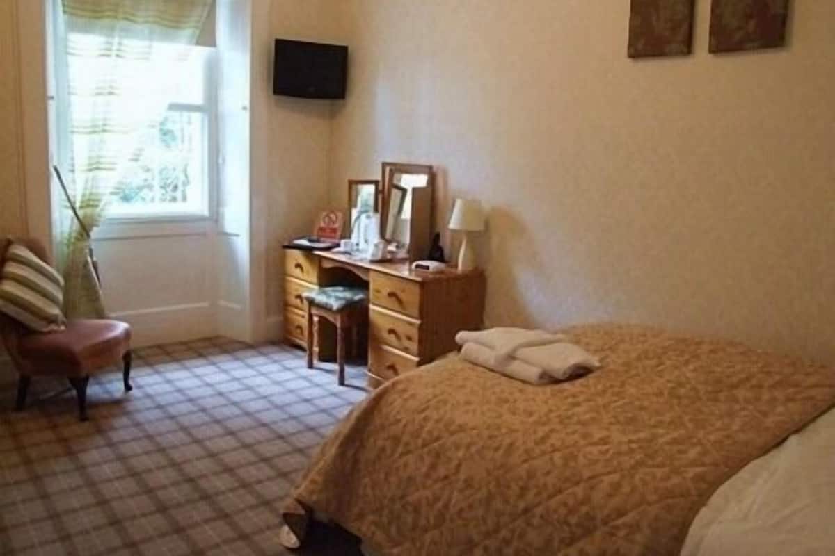 Quarto com cama, cadeira, escrivaninha, televisão e janela com cortinas.