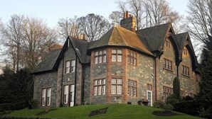 A large, stone building with multiple chimneys and a sloped roof, surrounded by trees and a stone wall. - The Glen Guesthouse (Selkirk)