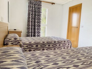 A bedroom with two beds, a wooden desk, a window with curtains, and a wooden door.