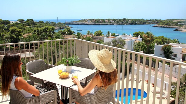 A balcony with two wicker chairs, a table with a plant and a fruit bowl, and a view of the sea and buildings. - Aparthotel Niu d´Aus (Santanyi)