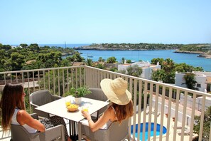 A balcony with two wicker chairs, a table with a plant and a fruit bowl, and a view of the sea and buildings.