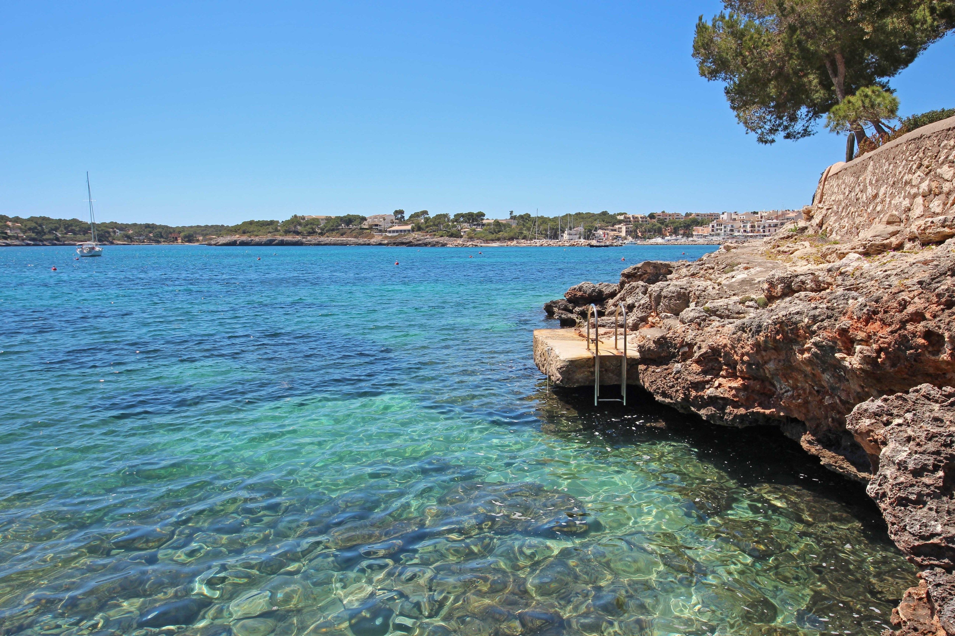 Paisaje costero con agua cristalina, costa rocosa y un velero a lo lejos.