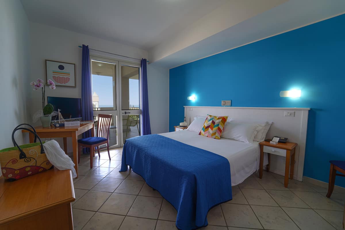 A hotel room with a bed, a desk, a chair, a small table, and a window with curtains.