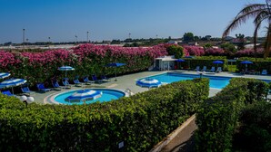 Un área de piscina con múltiples sillas de descanso y sombrillas, rodeada de exuberante follaje y arbustos florecientes de color rosa.