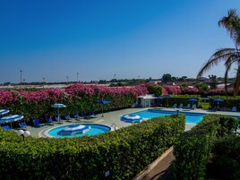 Un área de piscina con múltiples sillas de descanso y sombrillas, rodeada de exuberante follaje y arbustos florecientes de color rosa.