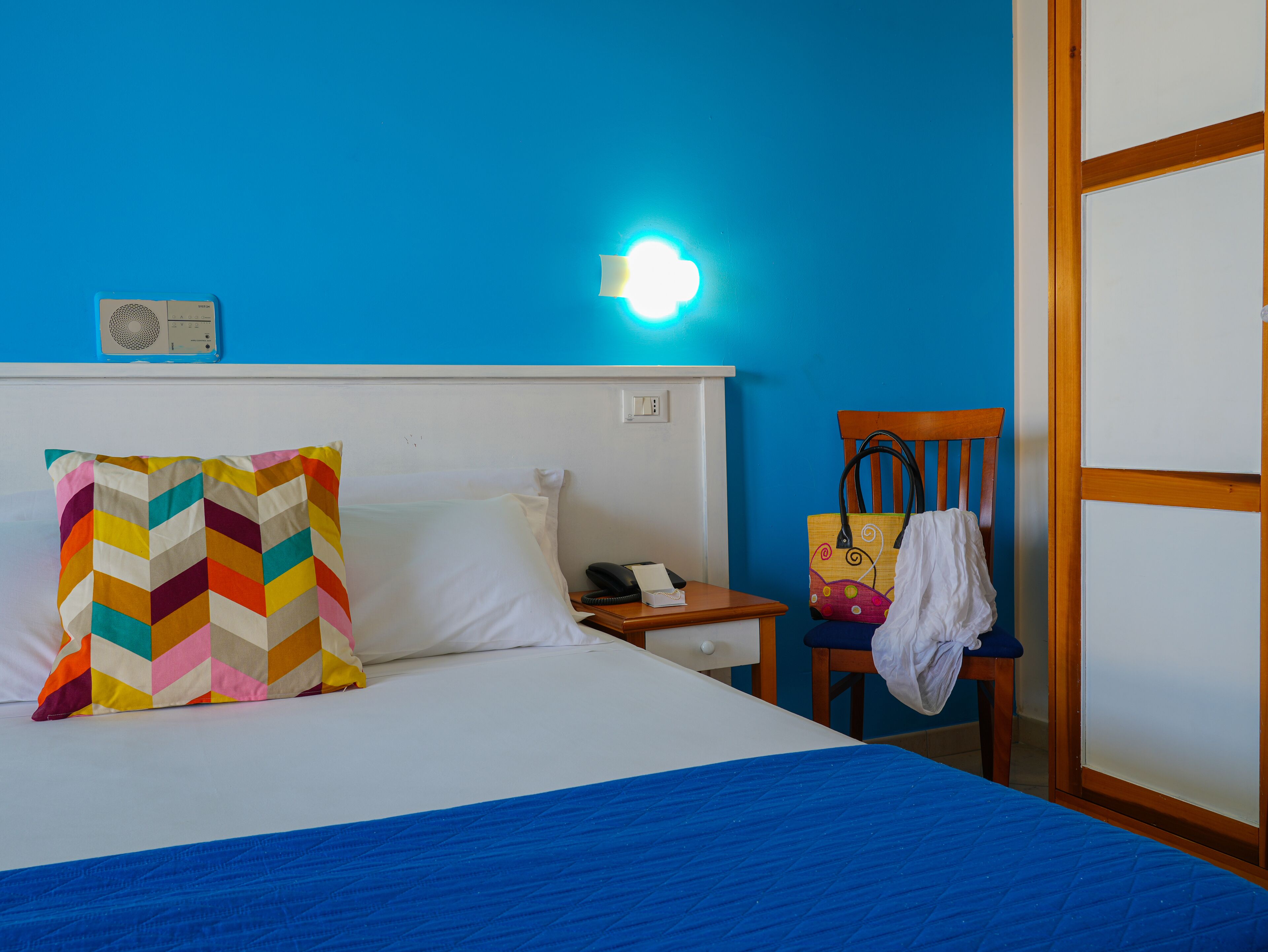 Una camera d'albergo con un letto, un comodino, una sedia e una lampada a parete.